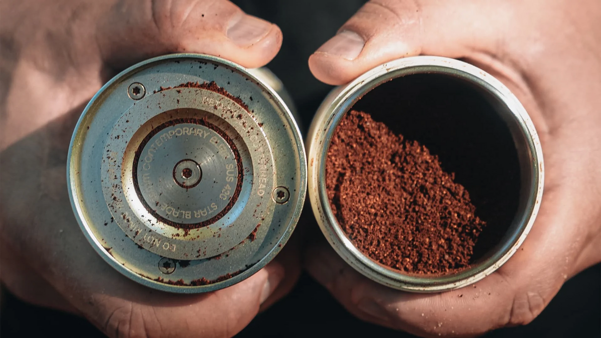 Nahaufnahme der Einzelteile einer manuellen Kaffeemühle mit frisch gemahlenem Kaffee in der Hand, wobei das Mahlwerkdesign und die Gleichmäßigkeit des Mahlguts hervorgehoben werden.