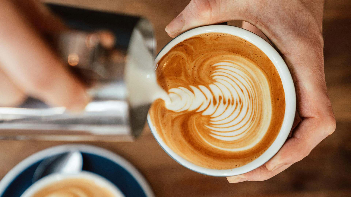 Nahaufnahme einer Hand, die aufgeschäumte Milch in ein Espressogetränk gießt und dabei gekonnt ein federleichtes Latte-Art-Muster in einer weißen Keramiktasse kreiert.