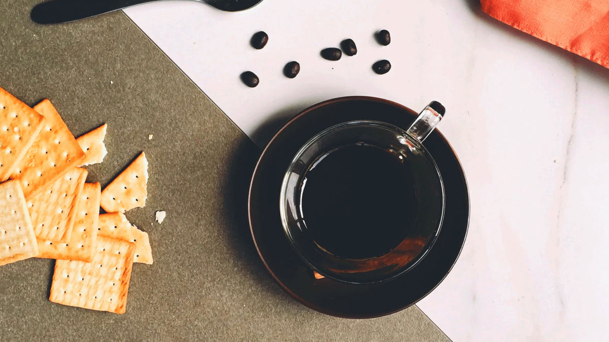 Espresso in Glastasse auf dunkler Untertasse, mit Kaffeebohnen und knusprigen Crackern auf hellem Tisch
