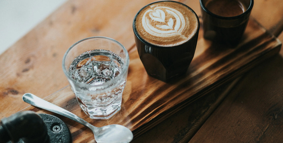 Latte Art mit Herz, dazu ein Glas Wasser und Löffel auf einem Holztablett