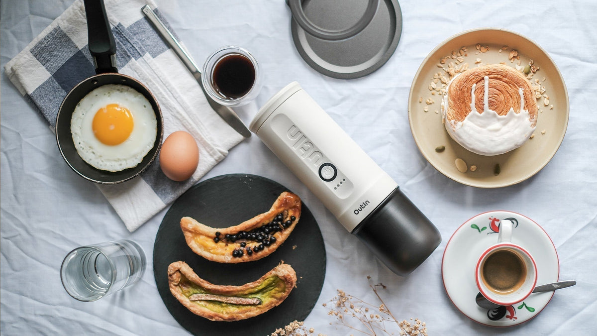Frühstücksszene mit Outin tragbarer Espressomaschine, Spiegelei, Gebäck und Kaffeetassen auf weißer Tischdecke, die zeigt, wie Kaffeegenuss mit dem Frühstück für optimale Verdauungsvorteile kombiniert wird.
