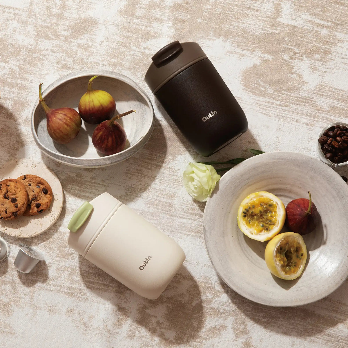 Zwei OutIn Reisebecher in Dunkelbraun und Cremeweiß mit Lebensmitteln wie Feigen, Passionsfrucht und Kaffeebohnen.