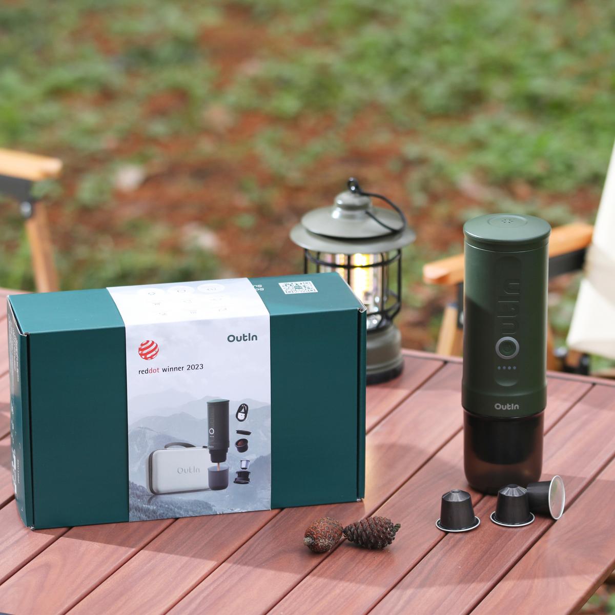 Waldgrüner OutIn Reise-Espressomaschine mit Verpackung, Red Dot Award und Kaffeekapseln im Freien.
