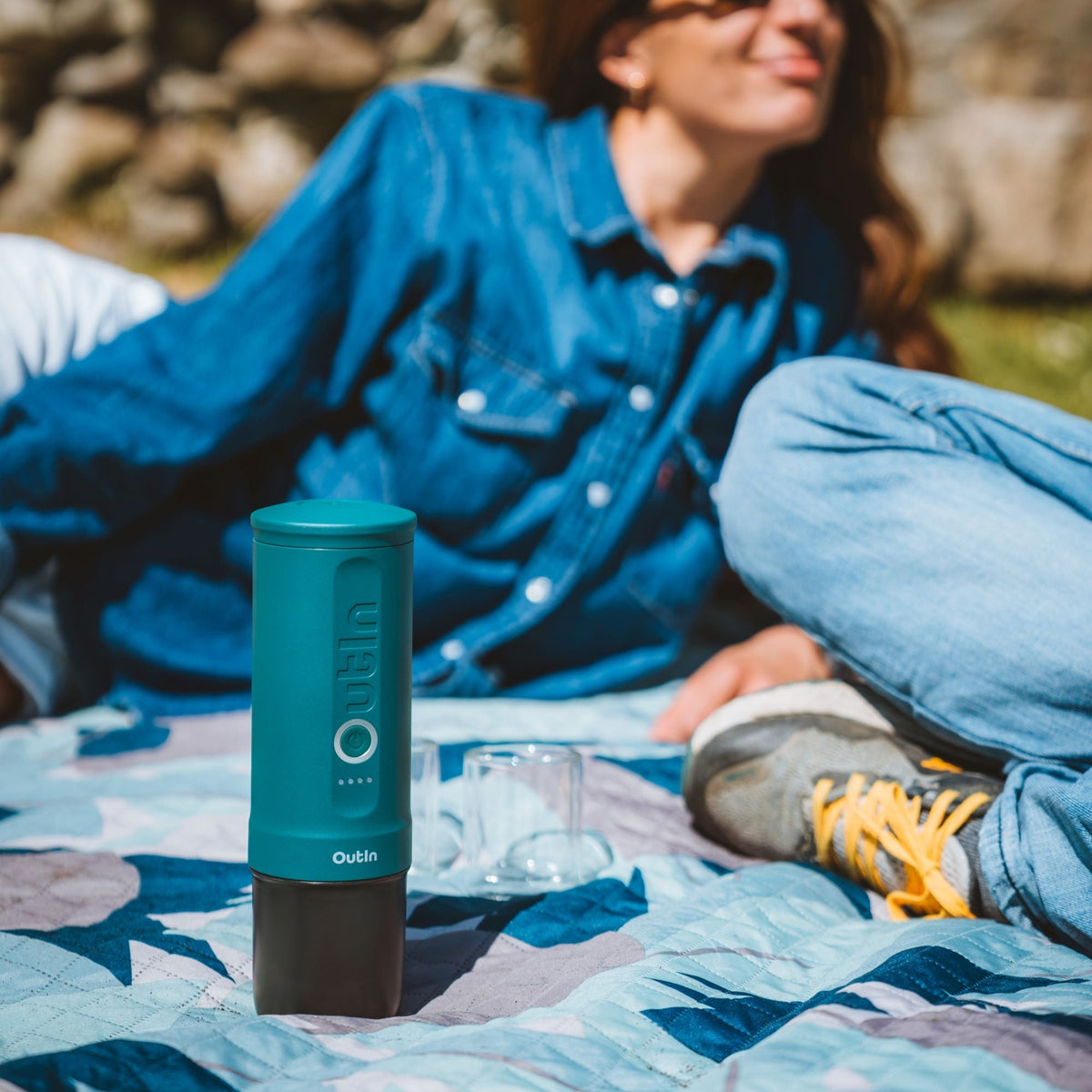 Nano Tragbare Espressomaschine Blaugrün auf Picknickdecke mit Person im Hintergrund, ideal für Outdoor-Kaffee.