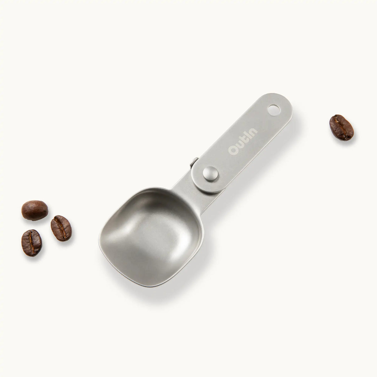 OutIn faltbarer Kaffeelöffel aus Edelstahl mit Aufhängeloch und Kaffeebohnen auf weißem Hintergrund.
