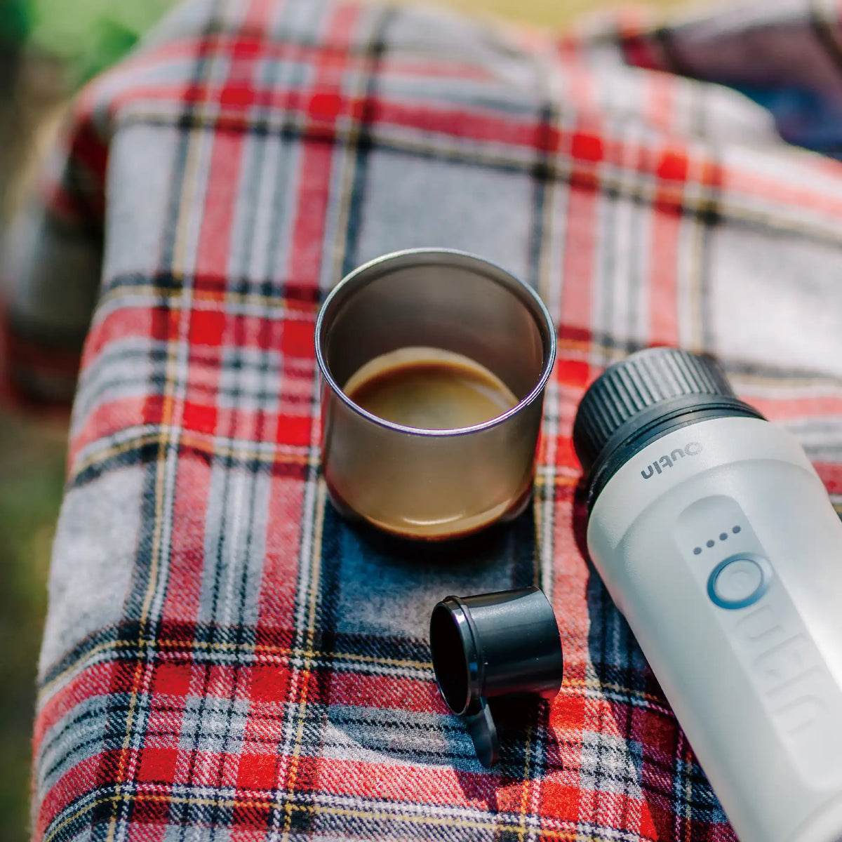 Edelstahlbecher mit Espresso neben Nano Espressomaschine auf karierter Decke für Outdoor-Kaffee.