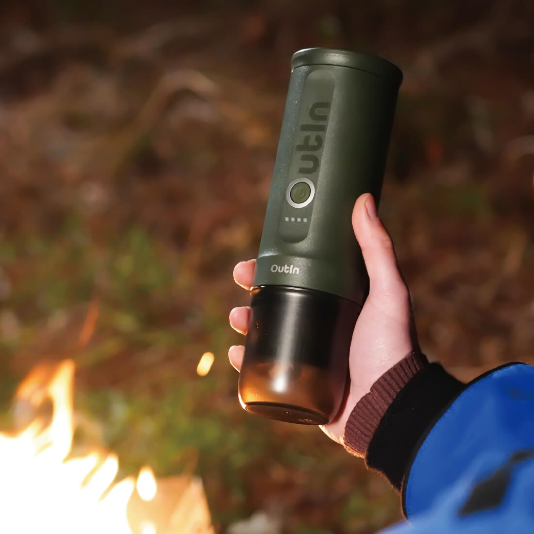 Waldgrüne OutIn Nano Espressomaschine in der Hand vor Lagerfeuer, für Camping und Outdoor geeignet.