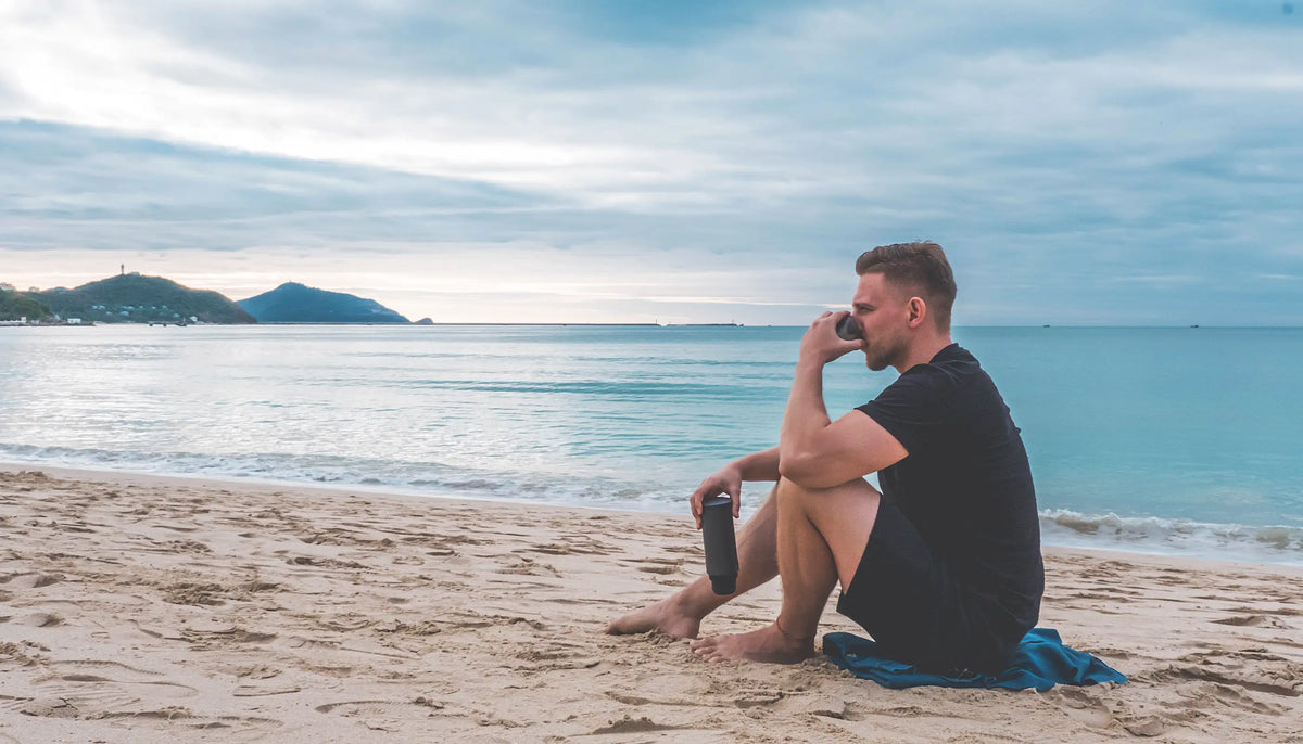 Mann entspannt am Strand mit Outin-Kaffeebereiter, genießt den Meerblick bei Sonnenaufgang, Outdoor-Kaffeegenuss am Meer﻿