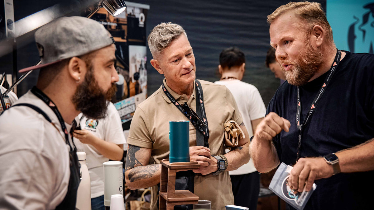 Gespräch unter Kaffeefachleuten am Messestand: Vorführung eines Brühgeräts, Austausch zu Espresso‑Technik, Mühle und Zubehör.﻿
