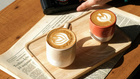 Eine gemütliche Kaffeepause: Zwei Lattes mit Latte Art, serviert auf einem Holztablett und einer Vintage-Zeitung im warmen Sonnenlicht