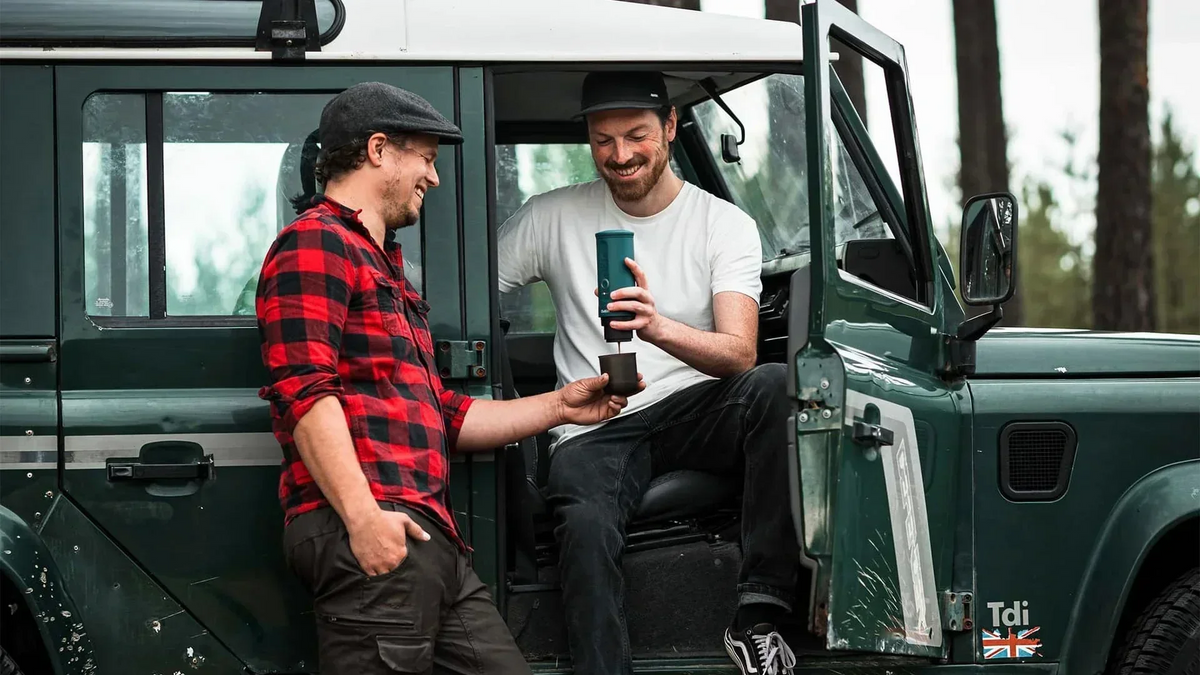 Zwei Personen genießen Kaffee im Freien neben einem grünen Geländewagen mit geöffneter Tür