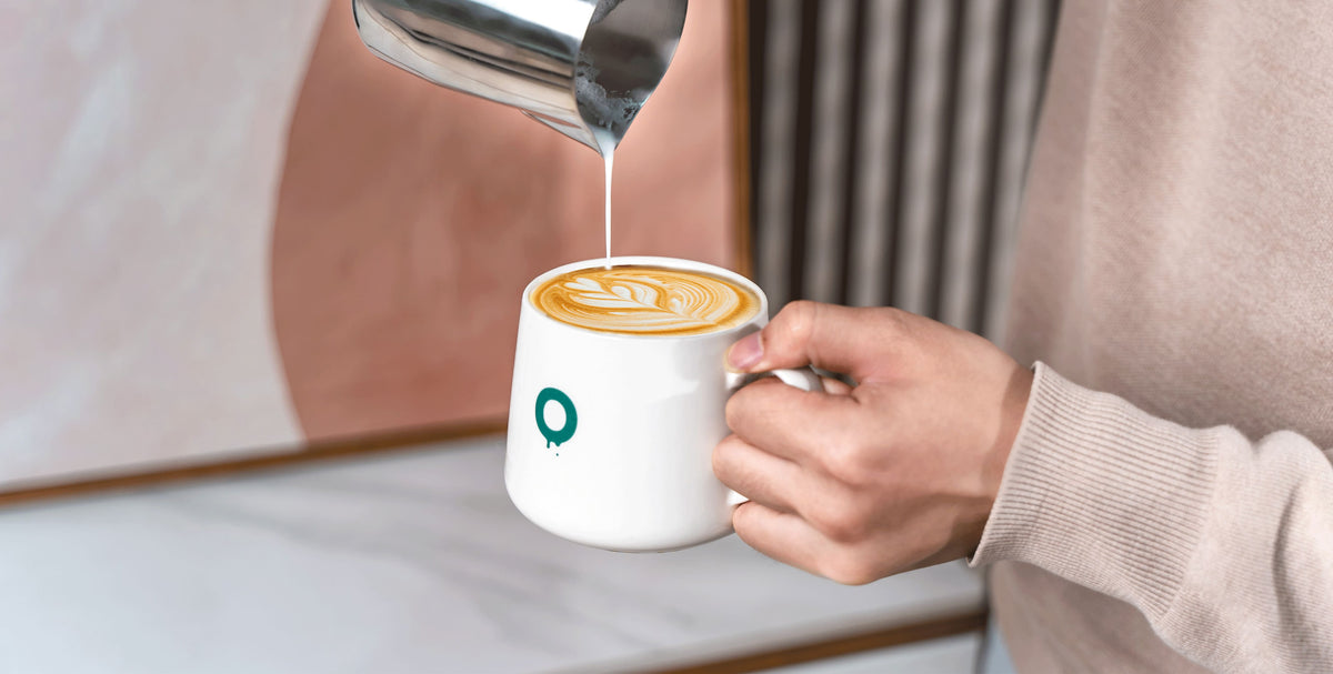 Ein Barista gießt geschäumte Milch in einen Latte in einer weißen Tasse mit dem O-Logo darauf.