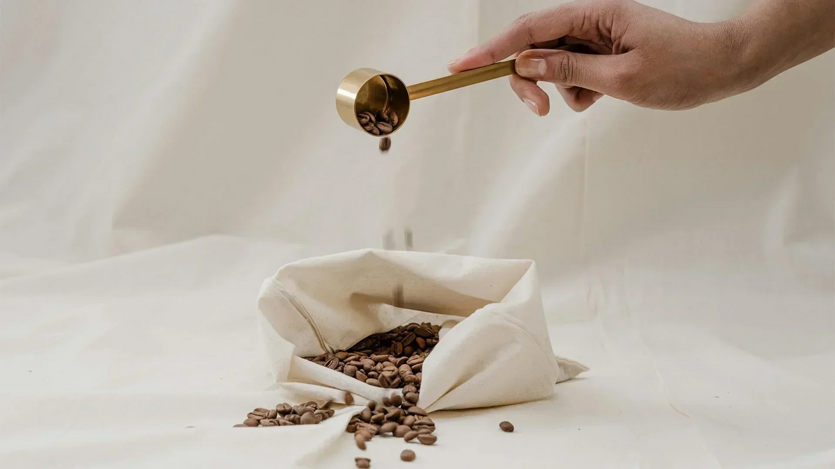 Kaffeebohnen werden mit einem goldenen Messlöffel in einen hellen Stoffbeutel gefüllt.