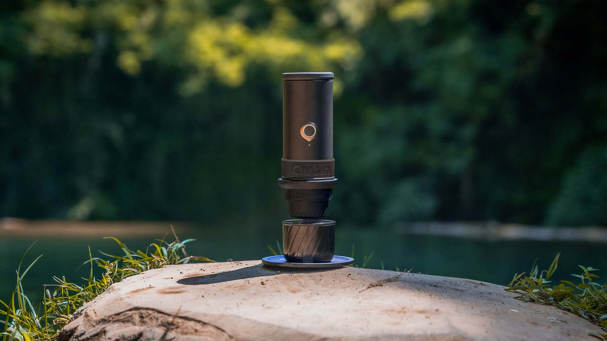 Eine tragbare Espressomaschine, die gerade einen Espresso in eine Tasse brüht, steht auf einem hölzernen Baumstumpf an einem See.