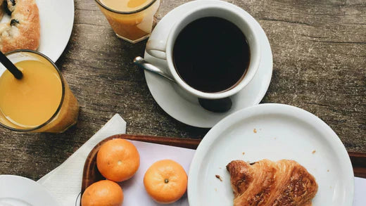 A breakfast scene with a cup of black coffee, fresh croissants and whole oranges on white plates atop a rustic wooden table.