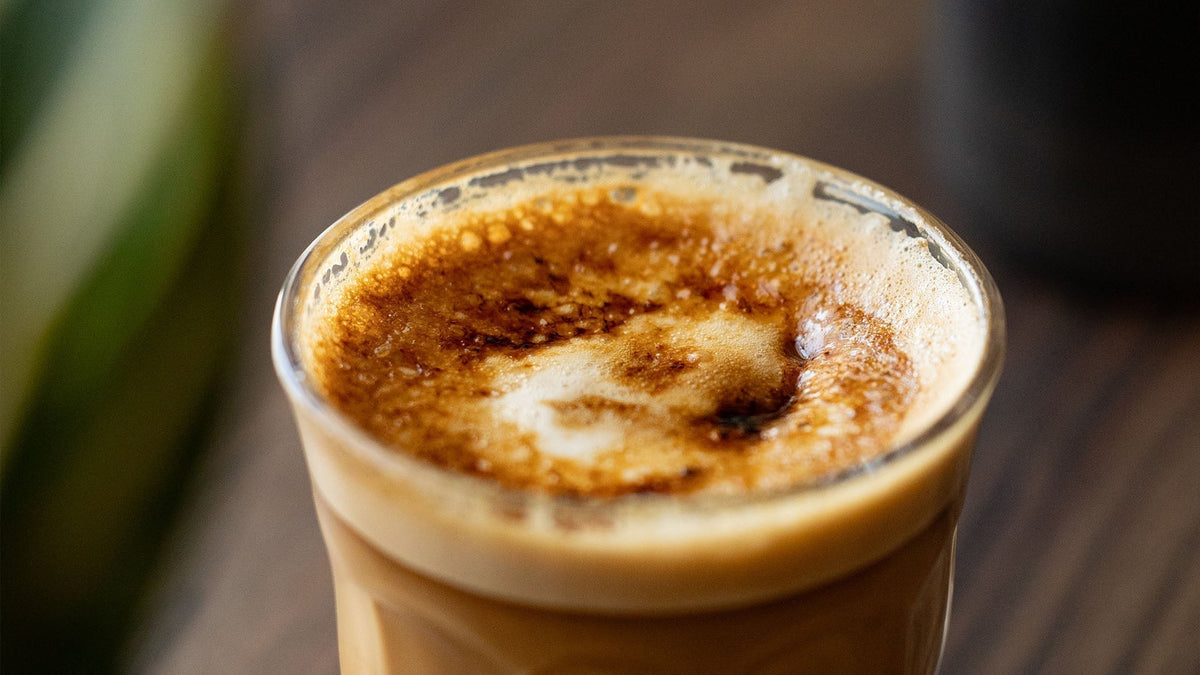 Nahaufnahme von frisch gebrühtem Kaffee in einem Glas, der eine reichhaltige goldbraune Crema mit kleinen Bläschen auf der Oberfläche zeigt und die perfekte Grundlage für Latte-Art-Designs bietet.
