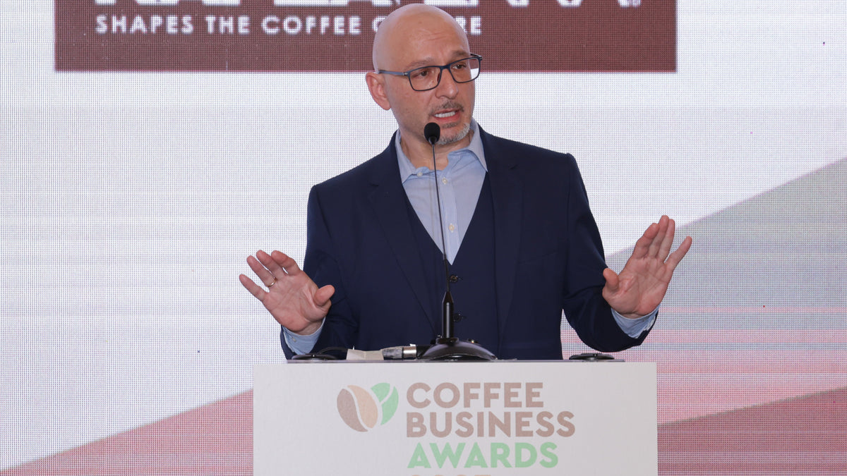 Panagiotis hält eine Rede bei den Coffee Business Awards. Der Mann im Anzug gestikuliert vor der Leinwand mit Kaffeemotto