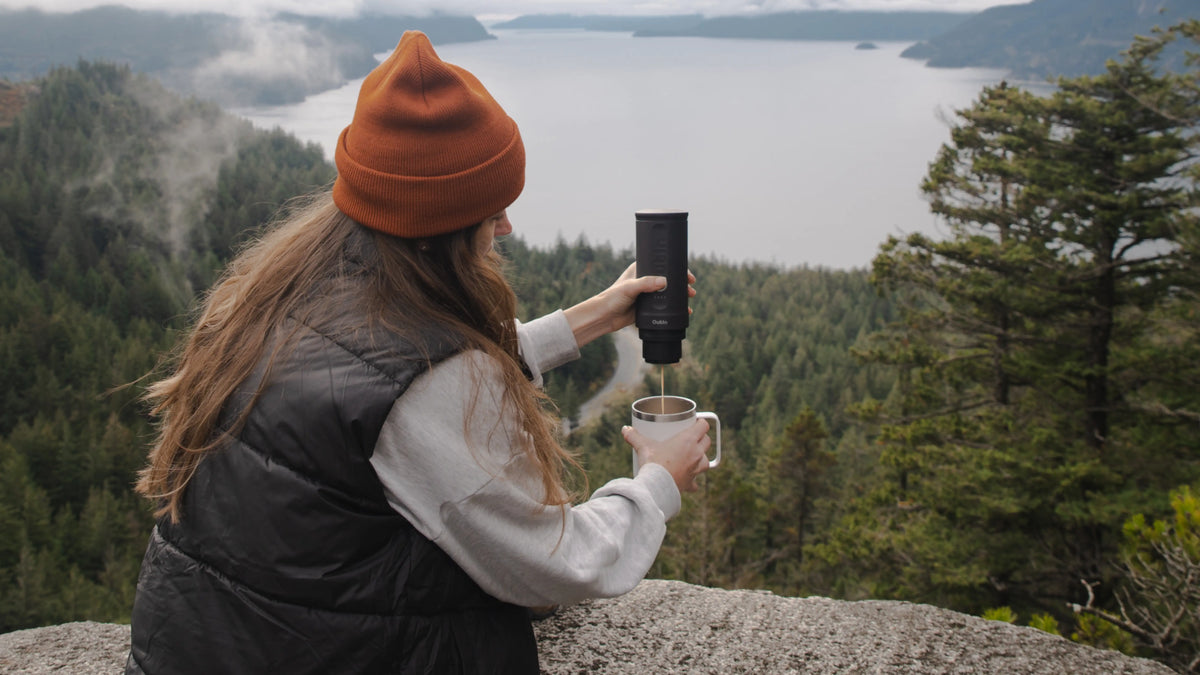 Eine Person brüht frischen Kaffee mit einer tragbaren Espressomaschine vor der Kulisse eines malerischen Bergsees und demonstriert so die bequeme Kaffeezubereitung inmitten der Natur.