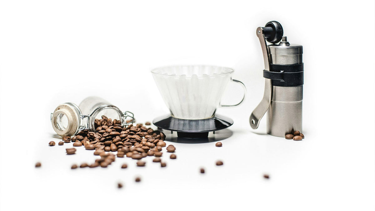 Komplettes Pour-Over-Kaffeeset mit Handkaffeemühle, Glasfilter und frischen Kaffeebohnen auf weißem Hintergrund – minimalistisches Arrangement von Kaffeezubehör.