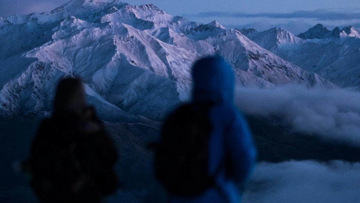 Zwei Personen in Winterkleidung betrachten unscharf schneebedeckte Berge bei Dämmerung. Klare Berglandschaft im Hintergrund.