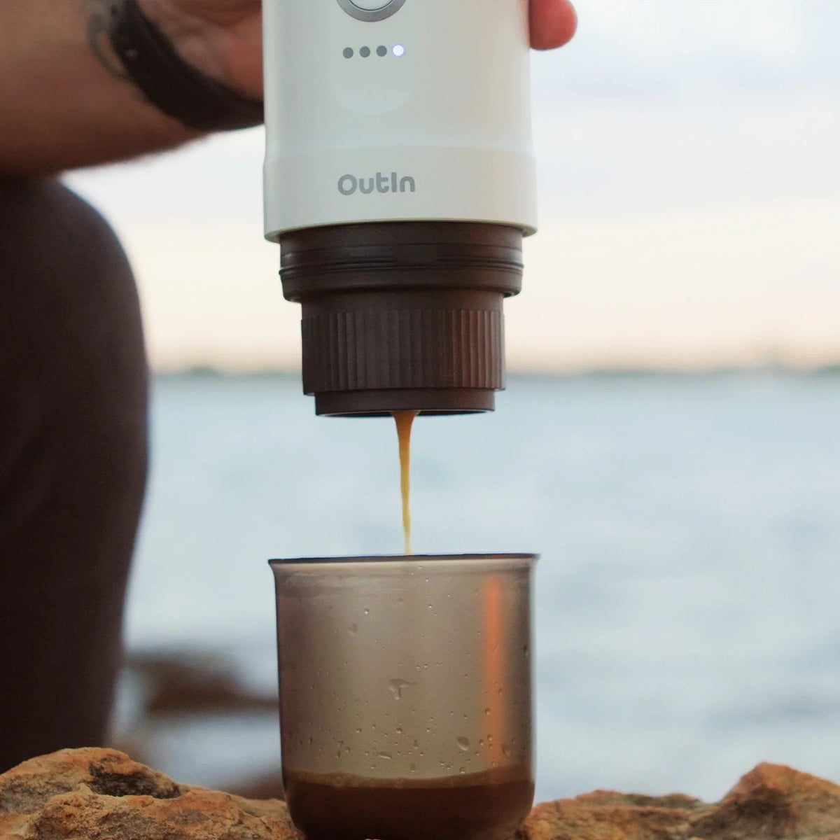 Perlweiße OutIn Nano Espressomaschine brüht draußen Espresso in einen grauen Becher auf Felsen am Wasser.