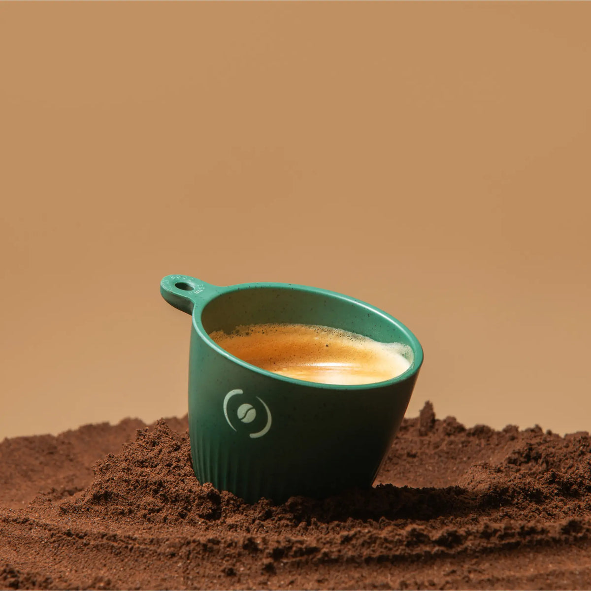 Tiefgrüner OutIn Rebrew Espressobecher mit Henkel, gefüllt mit cremigem Espresso, steht auf Kaffeepulver.