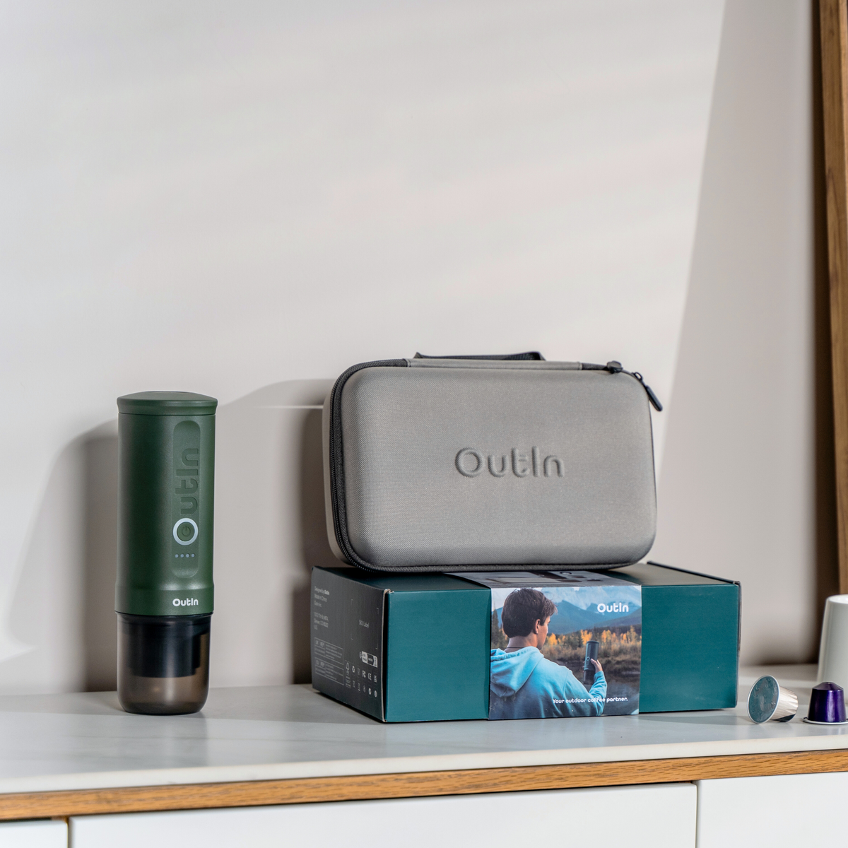 Waldgrüne OutIn Reise-Espressomaschine mit Zubehör: Schutzhülle, Verpackung & Kaffeekapseln auf dem Tisch.