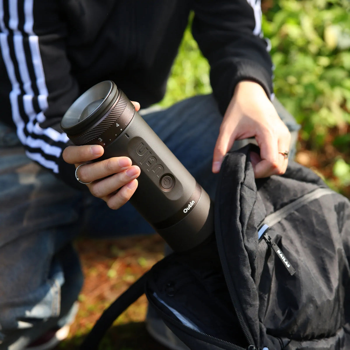 Person packt OutIn Fino tragbare Kaffeemühle in schwarzen Rucksack, ideal für Reisen. Mahlgradskala sichtbar.