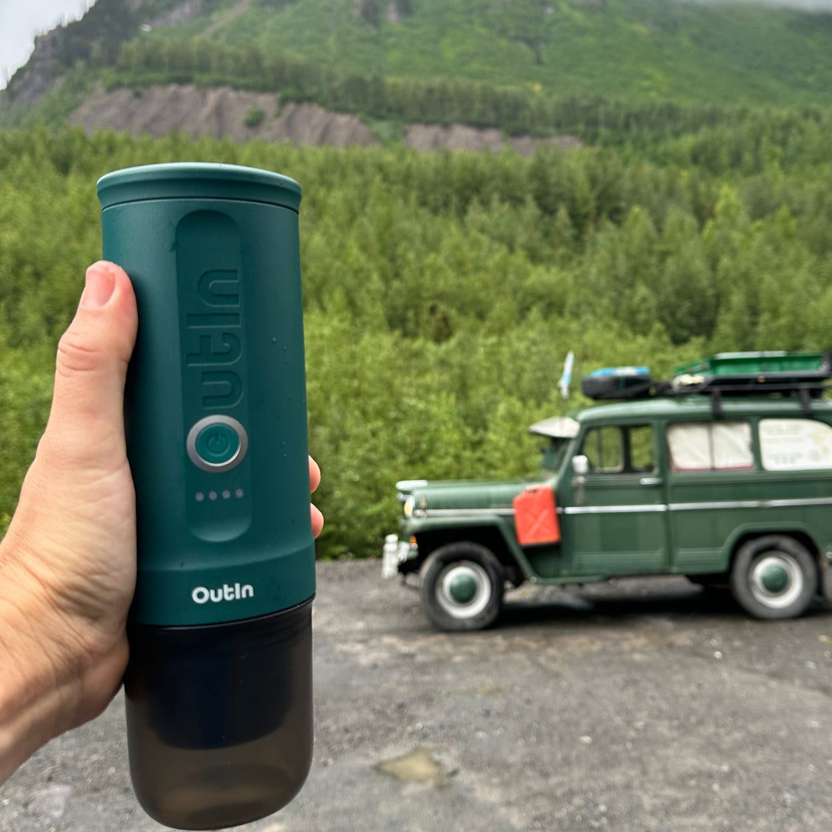 Nano tragbare Espressomaschine blaugrün von Hand gehalten vor einem Oldtimer und Berglandschaft. Ideal für Outdoor.